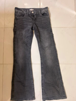 Grå bootcut jeans med broderi - Snygga grå bootcut jeans med broderade detaljer på bakfickorna. Jeansen har klassisk femficksmodell, knappgylf och utsvängda ben. Materialet är denim i bomull med en tvättad look. Perfekta för dig som vill ha en unik touch på din outfit.
