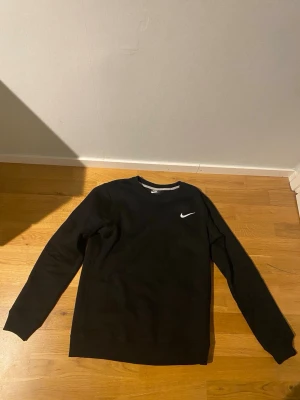 Svart sweatshirt från Nike - Klassisk svart sweatshirt från Nike med rund halsringning och vit broderad Swoosh-logga på bröstet. Tröjan har långa ärmar och ribbade muddar vid ärmslut och nederkant. Perfekt för en sportig och avslappnad stil.