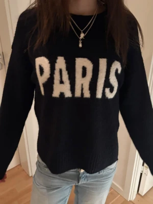 Svart stickad tröja med PARIS-tryck - Cool svart stickad tröja med stort vitt PARIS-tryck på bröstet. Tröjan har en rak, något kortare passform och långa ärmar. Perfekt för dig som gillar streetstyle och vill ha en statement-piece i garderoben.