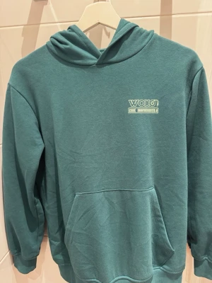 Grön hoodie från Woxo 720 - Snygg turkos hoodie från Woxo med tryck på bröstet och stor känguruficka framtill. Tillverkad i mjukt och bekvämt material, perfekt för chill dagar. Klassisk huva och ribbade muddar vid ärmar och nederkant.