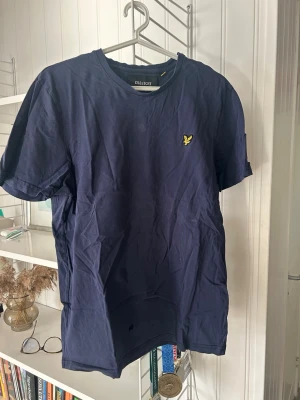 Marinblå t-shirt från Lyle & Scott - Snygg marinblå t-shirt från Lyle & Scott med klassisk gul logga på bröstet. T-shirten har rund halsringning och korta ärmar. Tillverkad i mjuk bomull som känns skön mot huden. Perfekt för en clean och stilren look.