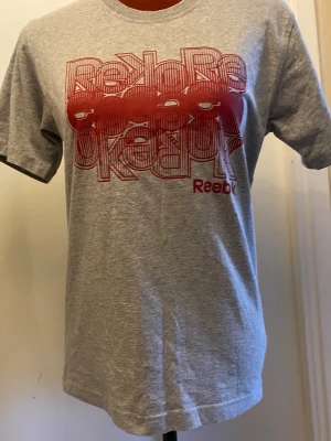 Grå Reebok t-shirt med röd print XS - Snygg grå t-shirt från Reebok med ett stort rött grafiskt tryck och logga på bröstet. Klassisk passform och rund halsringning. Perfekt för en sportig och avslappnad stil.