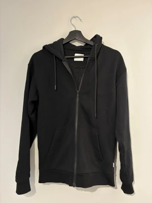 Svart zip hoodie med huva - Klassisk svart hoodie med dragkedja framtill och stor ficka på magen. Modellen har huva med dragsko och ribbade muddar vid ärmar och nederkant. Perfekt för en avslappnad och stilren look. Materialet känns mjukt och bekvämt, troligen bomullsblandning.
