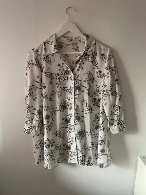 Vit blommig skjorta med knappar - Säljer en vit skjorta med svart och ljusblått blommönster. Skjortan har långa ärmar med knapp vid manschetten och klassisk krage. Med sömmar vid bröstet som ger en figursydd modell.