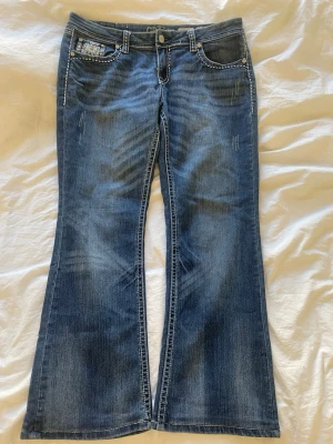 Blå bootcut jeans med broderade fickor - Säljer ett par blå bootcut jeans med snygga slitningar. Baksidan har två bakfickor med detaljerade broderier och nitar. Jeansen har låg midja och klassisk femficksdesign. Perfekta för dig som gillar en retro och unik stil.