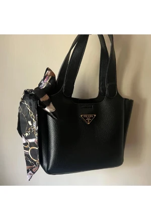 Svart handväska från Prada - Snygg svart handväska från Prada i strukturerat läder med dubbla handtag. Väskan har en dekorativ scarf i svart, lila och guldmönster knuten på sidan samt den klassiska Prada-loggan i metall framtill. Perfekt accessoar för att lyfta din outfit.