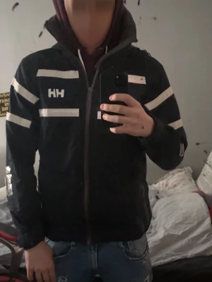 vind/seglarjacka från Helly Hansen - Snygg vindjacka från Helly Hansen med vita reflexdetaljer och dragkedja framtill. Jackan har hög krage, praktiska fickor och HH-logga på bröstet. Perfekt för blåsiga dagar och outdoor-aktiviteter. Tveka inte att skriva. Fraktar samma dag.