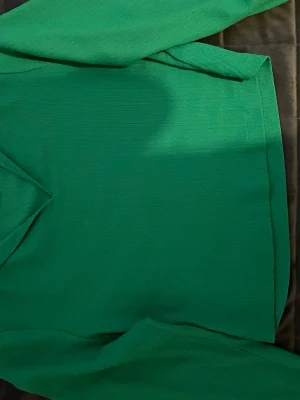 Grön långärmad blus - En stilren grön blus med långa, volang ärmar och rak passform. Tillverkad i ett lätt, strukturerat tyg som ger en modern känsla. Perfekt att matcha med jeans eller kjol för en trendig look. Står S på blusen men passa även xs och xxs.