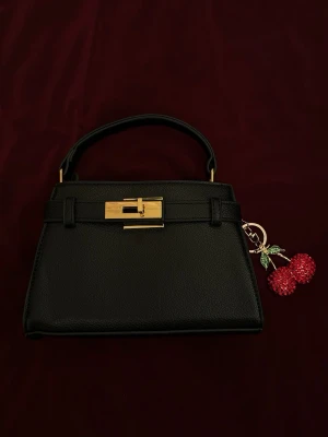 Svart handväska med körsbärsberlock - Stilren svart handväska i skinnimitation med guldfärgat spänne och kort handtag. Väskan har en dekorativ körsbärsberlock i rött och grönt som ger en lekfull touch. Perfekt accessoar för att lyfta din outfit.