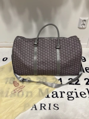 Grå Goyard resväska med axelrem - Snygg grå resväska från Goyard med det klassiska mönstret och detaljer i grått läder. Väskan har både handtag och justerbar axelrem för smidig användning. Perfekt för weekendresor och har en stilren, tidlös look. Levereras med original dustbag.