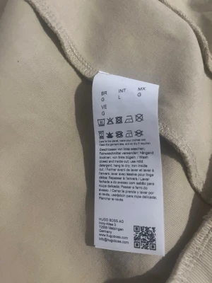 Beige hoodie från BOSS - Snygg beige hoodie från BOSS i mjukt material. Klassisk design med ribbade muddar och diskret logga. Perfekt för en avslappnad och stilren look. Passar till det mesta och är enkel att matcha med andra plagg.