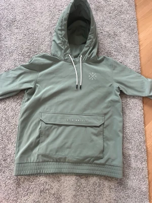 Grön hoodie från Dope med magficka - Snygg ljusgrön hoodie från Dope med stor magficka och vit dragsko i huvan. Tryck med Dope-logga på bröstet och text på ficklocket. Perfekt för dig som gillar streetwear och vill ha en stilren look.