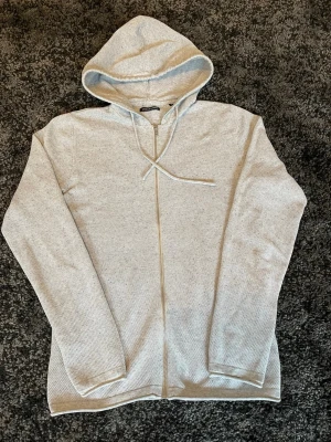 Ljusgrå zip-hoodie från Jack&Jones  - Ljusgrå hoodie med dragkedja från Jack&Jones. Tröjan har huva med dragsko och är tillverkad i ett mjukt, strukturerat tyg. Perfekt för en avslappnad och stilren look. Långa ärmar och enkel design utan tryck eller mönster.