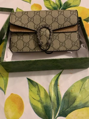 Gucci Dionysus GG Supreme handväska - Säljer en beige och brun Gucci Dionysus handväska i GG Supreme canvas med ikoniskt monogrammönster och silverfärgat spänne med ormdetaljer. Klassisk rektangulär form och lockstängning. Perfekt för dig som vill ha en statementväska från Gucci.