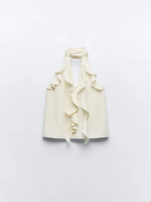 Krämvit volangblus med hög krage - Stilren krämvit blus med volangdetaljer framtill och hög krage. Blusen är ärmlös och har en elegant, lös passform. Perfekt för dig som gillar en chic och trendig look. Materialet ser ut att vara ett mjukt, lätt tyg som faller snyggt.