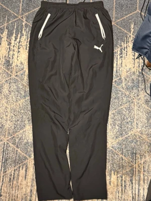 Svarta träningsbyxor från Puma - Säljer ett par svarta träningsbyxor från Puma med vita dragkedjor vid fickorna och bensluten. Byxorna har elastisk midja och är tillverkade i ett lätt syntetmaterial, perfekta för träning eller sportaktiviteter.