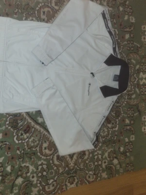 Vit vindjacka från Champion - Snygg vit vindjacka från Champion med svart krage och diskret logga på bröstet. Jackan har dragkedja framtill och långa ärmar. Perfekt för dig som vill ha en stilren och sportig look.