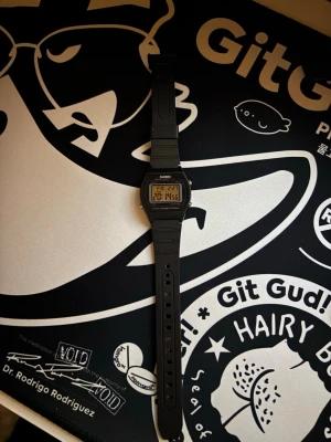 Svart digital klocka från Casio - Klassisk digital klocka från Casio i svart med rektangulär display och gummiband. Klockan har tydlig LCD-skärm, bakgrundsbelysning och flera funktioner som datum och alarm. Perfekt för dig som gillar retrostil och enkelhet.