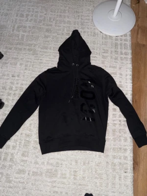 Svart hoodie från Dsquared2 ICON - Säljer en svart hoodie från Dsquared2 med stor glansig 'ICON'-text och logga på framsidan. Hoodien har dragsko i huvan och ribbade muddar vid ärmslut och nederkant. Perfekt för dig som gillar streetwear och vill sticka ut.