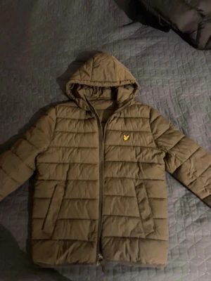 Olivgrön dunjacka från Lyle & Scott - Säljer en olivgrön dunjacka från Lyle & Scott med huva och dragkedja framtill. Jackan har det klassiska gula logomärket på bröstet, två sidofickor och quiltad design. Perfekt för kalla dagar och riktigt skön att bära.