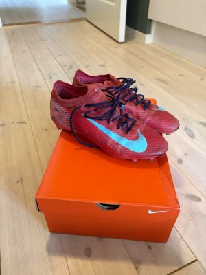 Nike röda fotbollsskor med blå swoosh - Säljer ett par röda Nike fotbollsskor med blå swoosh och svarta skosnören. Skorna har låg profil och är tillverkade i ett lätt syntetmaterial för bra bollkontroll. Perfekta för dig som vill ha stilrena och snabba skor på planen.