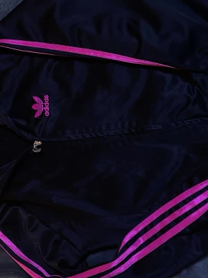 Svart Adidas ziptröja med rosa ränder - Snygg svart ziptröja från Adidas med klassiska rosa ränder längs ärmarna och rosa Adidas-logga på bröstet. Den är perfekt för en sportig fit. Det är bara att fråga om det är nått. 💕