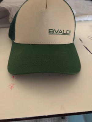 Grön och beige truckerkeps från BVALDI - Snygg truckerkeps från BVALDI i grön och beige med broderad logga framtill. Kepsen har klassisk böjd skärm, mesh baktill för extra ventilation och justerbar snapback. Perfekt för en avslappnad och trendig stil.