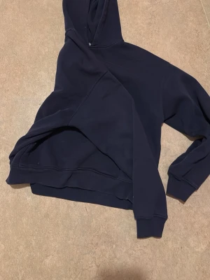 Mörkblå hoodie med huva - En stilren mörkblå hoodie med klassisk huva och ribbade muddar vid ärmslut och nederkant. Hoodien har en enkel design utan tryck eller mönster och är tillverkad i ett mjukt och bekvämt material, perfekt för en avslappnad look.