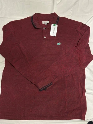 Vinröd långärmad skjorta från Lacoste - Säljer en vinröd långärmad skjorta från Lacoste i slim fit-modell. Skjortan har klassisk krage, knäppning framtill och Lacostes ikoniska gröna krokodillogga på bröstet. Materialet är mjukt och bekvämt, perfekt för en stilren look. Pris kan diskuteras kan passa för XL