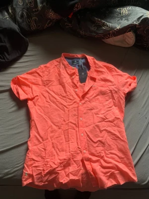 Korallfärgad skjorta från NZA - Kortärmad skjorta från NZA New Zealand Auckland i en snygg korallfärg. Skjortan har klassisk krage, knappar framtill och är tillverkad i bomull. Perfekt för dig som vill sticka ut med färg i sommar.