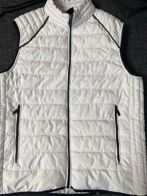 Vit dunväst med svarta detaljer från Newyorker  - Snygg vit dunväst med svarta dragkedjor och detaljer. Västen har ståkrage, två fickor med dragkedja och quiltad design. Perfekt för lager-på-lager och ger en clean look. Materialet är lätt och isolerande, perfekt för kyliga dagar.