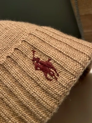 Beige mössa från Polo Ralph Lauren - Säljer en beige ribbstickad mössa från Polo Ralph Lauren med klassisk vinröd logga broderad framtill. Mössan är i mjukt stickat material och har vikt kant för extra stilpoäng. Perfekt accessoar för kyliga dagar.
