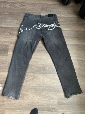 Svarta Ed Hardy jeans med tryck - Säljer ett par svarta jeans från Ed Hardy med stora vita logotryck bak och färgglada detaljer fram. Jeansen har normal passform och är gjorda i bomull. Perfekta för dig som gillar streetwear och vill sticka ut med coola prints.