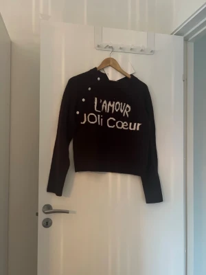 Svart stickad tröja med text - Cool svart stickad tröja med vit text 'L'AMOUR JOLI COEUR' framtill. Tröjan har asymmetrisk halsringning med dekorativa knappar på axeln och långa ärmar. Perfekt för dig som gillar statement-plagg och unik design.