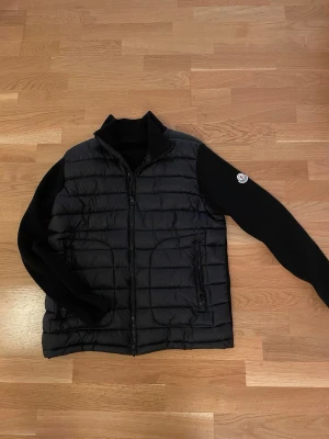 Svart cardiganjacka från Moncler - Snygg svart cardiganjacka från Moncler med vadderad framsida och stickade ärmar. Jackan har dragkedja framtill, två fickor med dragkedja och Moncler-logga på ena ärmen. Perfekt för lager-på-lager och streetstyle. Den har också nfc scan!