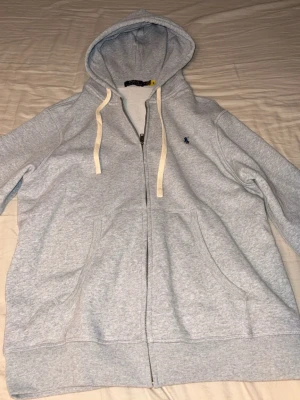 Ljusgrå hoodie från Polo Ralph Lauren - Ljusgrå zip-hoodie från Polo Ralph Lauren med vit dragsko i huvan och klassisk blå logga broderad på bröstet. Tillverkad i mjuk bomullsblandning och har två fickor framtill. Perfekt för en avslappnad och stilren look.
