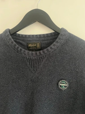 Mörkblå stickad tröja från Morris - Mörkblå stickad tröja från Morris med rund halsringning och ribbade muddar. Tröjan har en snygg patch med Morris-logga på bröstet och är tillverkad i ett mjukt, tjockt material som passar perfekt till kyligare dagar.