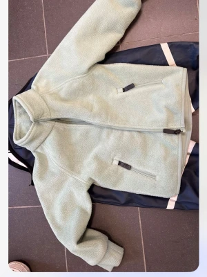 Ljusgrön fleecejacka från Kaxs - Mysig ljusgrön fleecejacka från Kaxs med dragkedja framtill och två praktiska fickor med dragkedja. Jackan har hög krage och ribbade muddar vid ärmsluten. Perfekt för lager på lager under kyliga dagar.