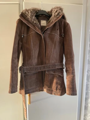 Brun mockajacka med fuskpäls och bälte - Säljer en brun mockajacka med huva fodrad i mjuk fuskpäls. Jackan har markerade sömmar, bälte i midjan och dragkedja framtill. Två stora fickor framtill och en skön passform. Perfekt för dig som vill ha en snygg och varm jacka med vintagekänsla. Står ingen storlek på den men skulle säga att den är som en s/m.
