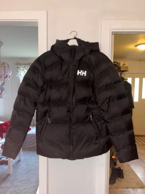 Svart pufferjacka från Helly Hansen - Säljer en svart pufferjacka från Helly Hansen med stor luva och dragkedja framtill. Jackan har två sidofickor med dragkedja och HH-logga på bröstet. Perfekt för kalla vinterdagar och riktigt skön att bära.
