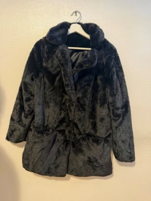 Svart kappa - Säljer en svart kappa i storlek L, den passar även som M. Jackan har en mjuk och fluffig yta, klassisk krage och stängs framtill. Tillverkad i 100% polyester med slätt innerfoder. Perfekt för att hålla värmen under kalla dagar och ger en trendig look.