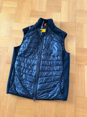 Svart dunväst från Parajumpers - Snygg svart dunväst från Parajumpers med quiltad framsida, hög krage och dragkedja. Västen har en gul detalj vid halsen och sidopaneler i stretchigt material för extra komfort. Perfekt för lager-på-lager och streetwear.