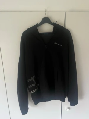Svart Champion hoodie med dragkedja - Säljer en svart hoodie från Champion med dragkedja framtill och huva med snören. Champion-logga på bröstet och coolt vitt mönster på sidan. Två fickor framtill. Perfekt för en chill och sportig look.