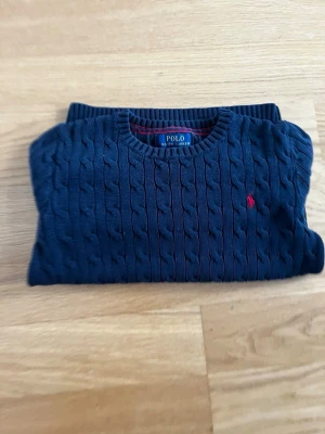 Mörkblå kabelstickad tröja Polo Ralph Lauren  - Mörkblå kabelstickad tröja från Polo Ralph Lauren med rund halsringning och röd broderad logga på bröstet. Tröjan har långa ärmar och ribbade muddar vid ärmslut och nederkant. Klassisk och tidlös design i mjukt material.