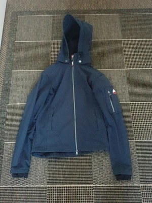 Mörkblå vindjacka från CRW XS - Snygg mörkblå vindjacka från CRW med huva och hel dragkedja framtill. Jackan har fickor med dragkedja och en extra ficka på ärmen. Perfekt för blåsiga dagar och har en sportig look med diskreta röda detaljer.