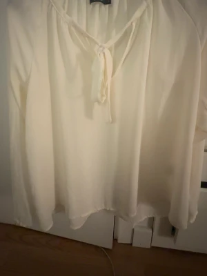 Vit blus med knytning från BikBok - Luftig vit blus från BikBok med långa ärmar och snygg knytning vid halsen. Tillverkad i ett tunt, lätt material som ger en avslappnad och stilren look. Perfekt att matcha med jeans eller kjol för en trendig outfit.