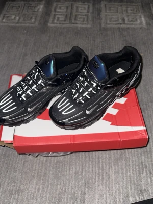 Nike Air Max Plus svarta sneakers - Nike Air Max Plus sneakers i svart med vita och silverfärgade detaljer. Skorna har en futuristisk design med synlig Air Max-dämpning i sulan och TN-logga på hälen. Ovandelen är i mesh och syntetmaterial med reflekterande detaljer och små Swoosh-loggor på sidorna.