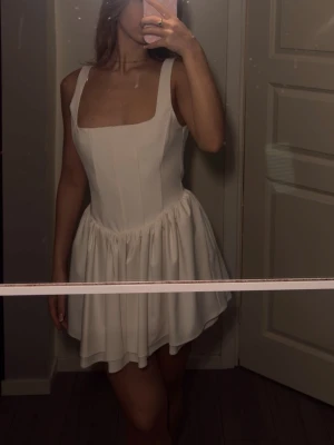 Vit kort klänning med breda axelband - Säljer en söt vit kort klänning med breda axelband och markerad midja. Klänningen har en voluminös kjol och fyrkantig urringning som ger en trendig och ungdomlig look. Perfekt för sommarens alla tillfällen! Helt oanvänd (etiketten sitter kvar ) 🥰