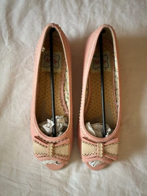 Rosa ballerinaskor med rosett  - Rosa ballerinaskor med krämfärgad rosett och detaljer💕 Liten klack  och öppen tå💕Originalförpackning kvar! Köpt secondhand men var tyvärr för stora så aldrig använda av mig😔Storlek 9, skulle säga att det är som storlek 40. Brukar ha 38/39 och de är för stora på mig!