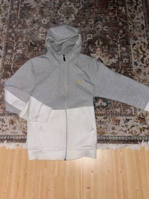 Gråvit hoodie med dragkedja - Snygg hoodie i tvåfärgad design med grå ovandel och vita partier nedtill och på ärmarna. Dragkedja framtill, huva med snören och ribbade muddar. Perfekt för en avslappnad streetwear-look.